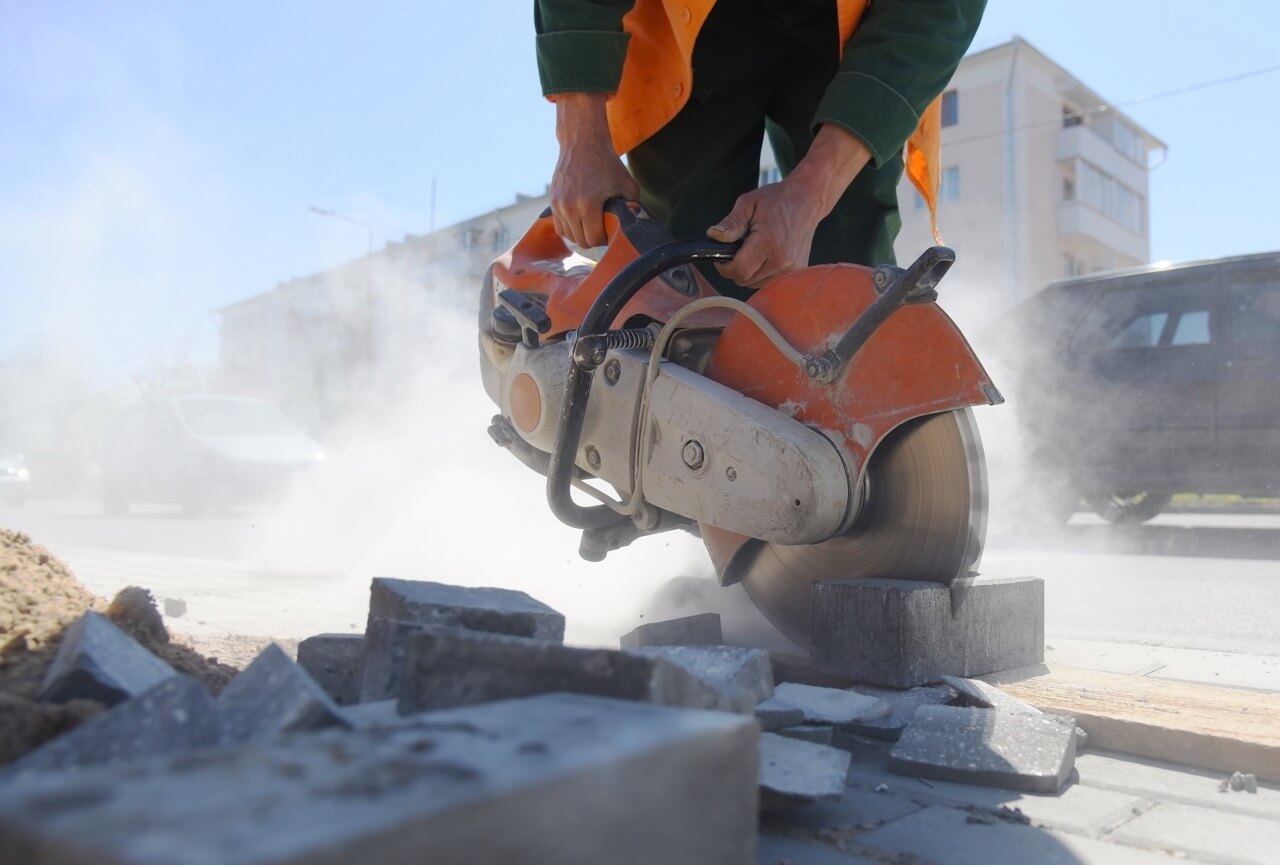 Worker using an industrial power saw to cut concrete blocks, creating airborne silica dust on a construction site, emphasizing the need for dust control and safety compliance.