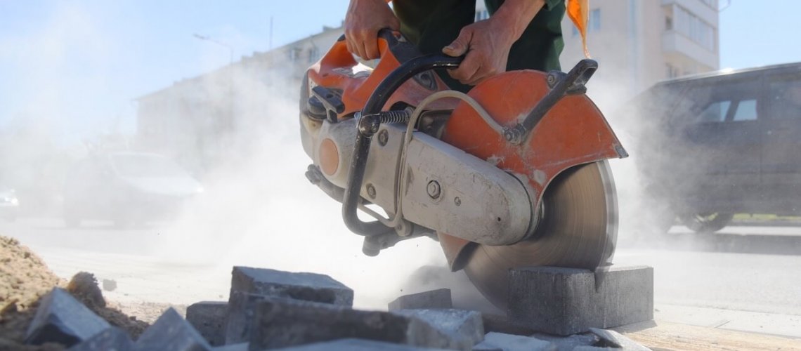 Worker using an industrial power saw to cut concrete blocks, creating airborne silica dust on a construction site, emphasizing the need for dust control and safety compliance.